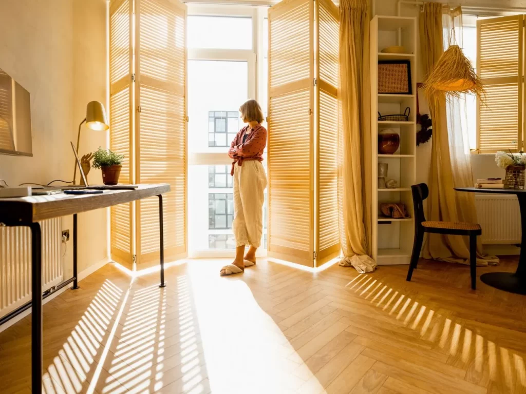 Donna appoggiata agli oscuranti della finestra che guarda la strada dalla finestra del salotto con il sole che entra e illumina la stanza